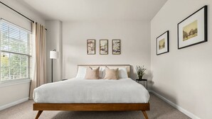 Ein Schlafzimmer mit Bett, einem Fenster mit Jalousien, einem Nachttisch und drei gerahmten Bildern an der Wand.