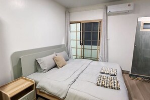 A single bed with a grey headboard, a bedside table with a drawer, a window with a view, and an air conditioning unit on the wall. - Saengcanthr (Nakhon Ratchasima)