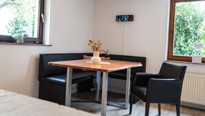 Una habitación pequeña y moderna con una mesa de madera, un sofá negro y una silla. Hay un reloj en la pared y una ventana con vistas a zonas verdes.