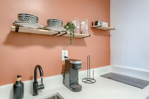 A kitchen with a white countertop, a coffee machine, a sink, and a wall-mounted shelf with dishes and a plant.