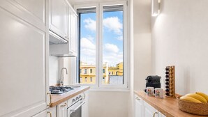 A narrow kitchen with white cabinetry, a gas stove, an oven, a window with a view of buildings, and a wooden countertop with a coffee maker and a fruit basket. - Michelangelo Prestige Apartment Vatican View by RHR (ROMA)