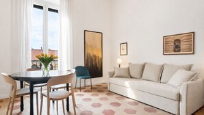 A modern living room with a round dining table, two chairs, a sofa, and a large window with curtains. - Michelangelo Prestige Apartment Vatican View by RHR (ROMA)