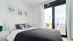 Ein modernes Schlafzimmer mit einem großen Bett, weißen Nachttischen und Blick auf die Stadt durch eine Glastür.