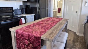 Una cocina con microondas, estufa y cafetera, una mesa de madera con mantel estampado de flores rojas, y una puerta que da a un baño.