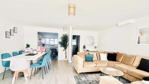Un soggiorno moderno con un divano angolare beige, una zona pranzo con sedie blu e una cucina sullo sfondo.