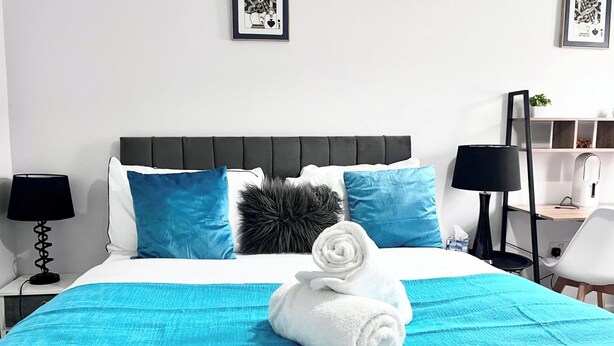 Un letto rifatto con una coperta blu, due cuscini, un asciugamano bianco arrotolato e un cuscino nero in pelo. Ci sono due lampade nere sui comodini, un tavolino con una lampada e una sedia.