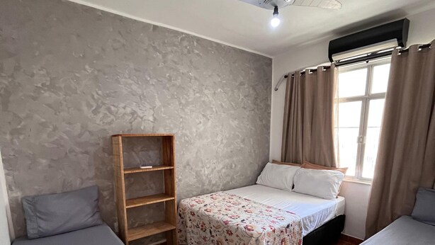 A room with two beds, a ceiling fan, a window with curtains, and a wooden shelf. - Colinna Hostel (Rio de Janeiro)