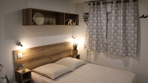 A bedroom with a wooden headboard, a bedside table with a lamp, a wall-mounted shelf with decorative items, and a patterned curtain. - Camping Charlieu - Nos Studios (Charlieu)