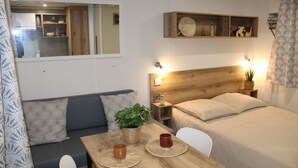 A compact bedroom with a wooden headboard, a bedside table with a lamp, a small shelf above the bed, a window with a view of the kitchen, and a sofa with cushions. - Camping Charlieu - Nos Studios (Charlieu)