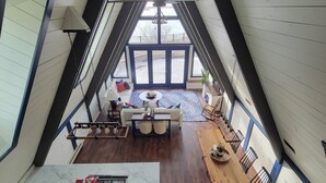 A high-ceilinged room with a sloped roof, a dining area with a wooden table and chairs, a sofa with cushions, and a view of the outdoors through large windows.