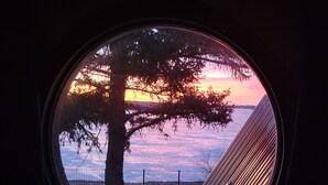 A window with a view of a sunset over a body of water, a tree, and a metal fence.
