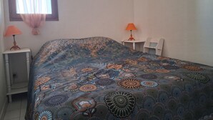 A bedroom with a patterned bedspread, two bedside lamps, a small white nightstand, and a window with sheer curtains.