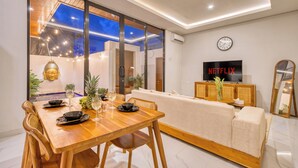 A modern living room with a wooden dining table set for two, a sofa, a television displaying "NETFLIX", and a large window with a view of the sky. - Cozy 3bedroom villa Bliss, Close to Canggu,
King bed,Tranquil! (Bali)