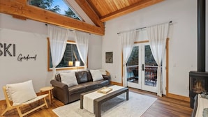 Un salon chaleureux avec un canapé, une table basse en bois et une cheminée. De grandes fenêtres sont ornées de rideaux, le plafond est en bois et un panneau est accroché au mur.