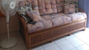 Una habitación con un sofá de mimbre, un ventilador de pie, un cuadro en la pared y una ventana con persianas.