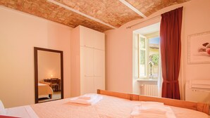 A hotel room with a large bed, a wooden headboard, a mirror, a window with curtains, and a radiator. - Apartment with garage - La Rosa nel Borgo (La Morra)