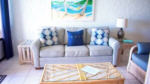 Ein Wohnzimmer mit einer Couch, einem Glastisch, einem Korbstuhl, einer Lampe und einem Gemälde von einer Strandansicht.