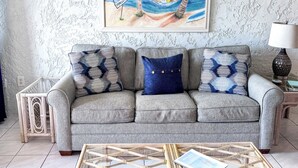 Ein Wohnzimmer mit einer grauen Couch, zwei gemusterten Kissen, einem Glastisch mit einem Notizbuch und einem Gemälde einer Strandansicht an der Wand.