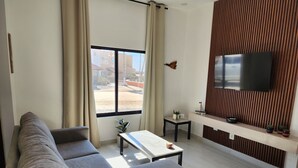 Ein modernes Wohnzimmer mit einem grauen Ecksofa, einem Couchtisch, einem an einer Holzvertäfelung befestigten Fernseher und einem kleinen Beistelltisch mit Dekoration.