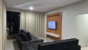 A modern living room with a sectional sofa, a flat-screen TV mounted on a wooden panel, and a small white shelf.