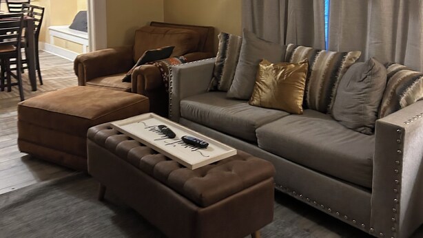 A living room with a sofa, ottoman, and a coffee table. There is a dining area visible in the background.