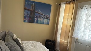 A bedroom with a bed, a nightstand, a door with curtains, and a painting of a bridge.