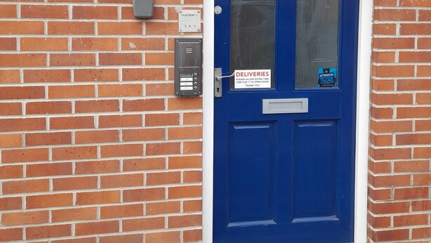 Eine blaue Tür mit Lieferhinweis, einem Briefkasten und einem Hausnummernschild an einer Backsteinmauer.