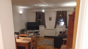 Ein Wohnzimmer mit Holzboden, einem Fernseher, einem Esstisch mit Stühlen, einer Couch und einer kleinen Kommode.