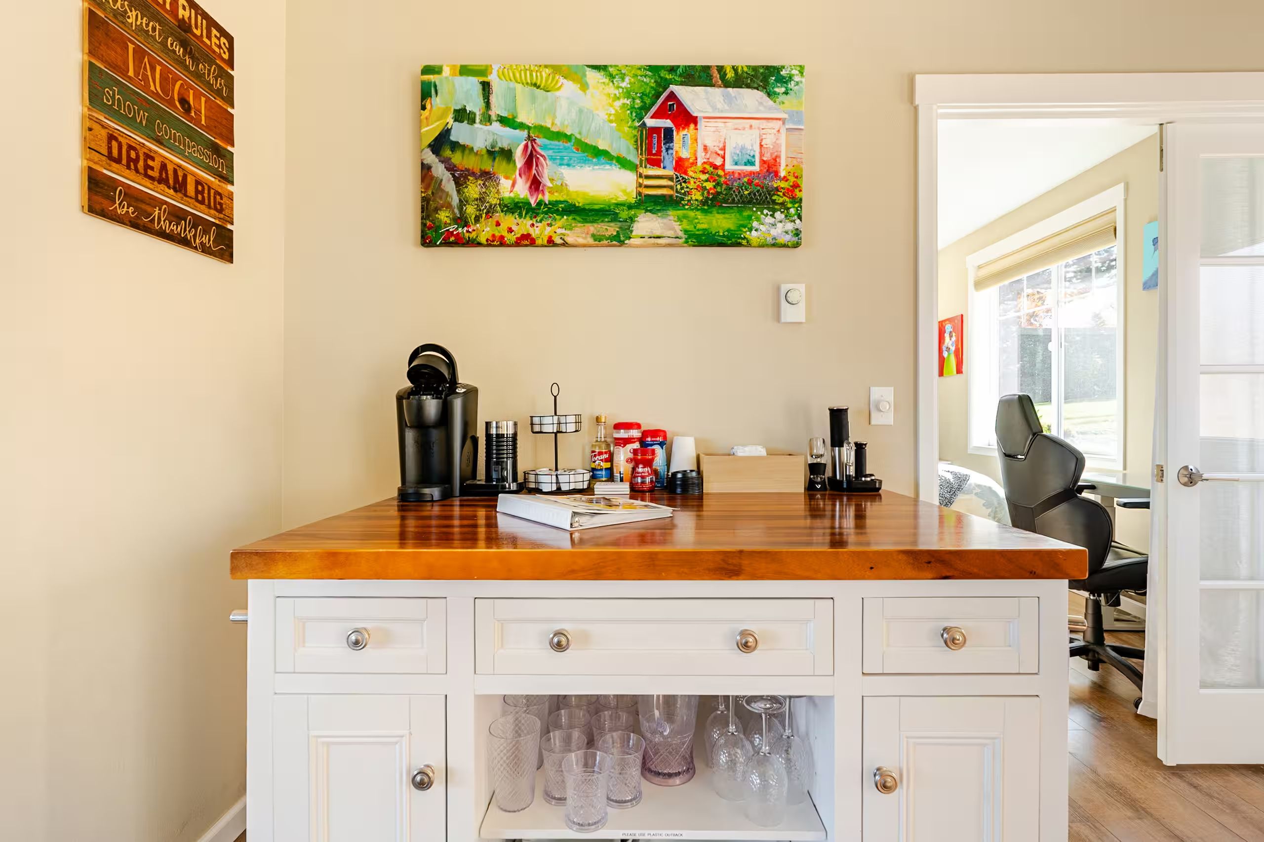A kitchen area with a wooden countertop, a coffee maker, and a painting of a house on the wall.
