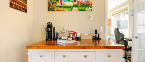 Un coin cuisine avec un plan de travail en bois, une machine à café et un tableau représentant une maison accroché au mur.
