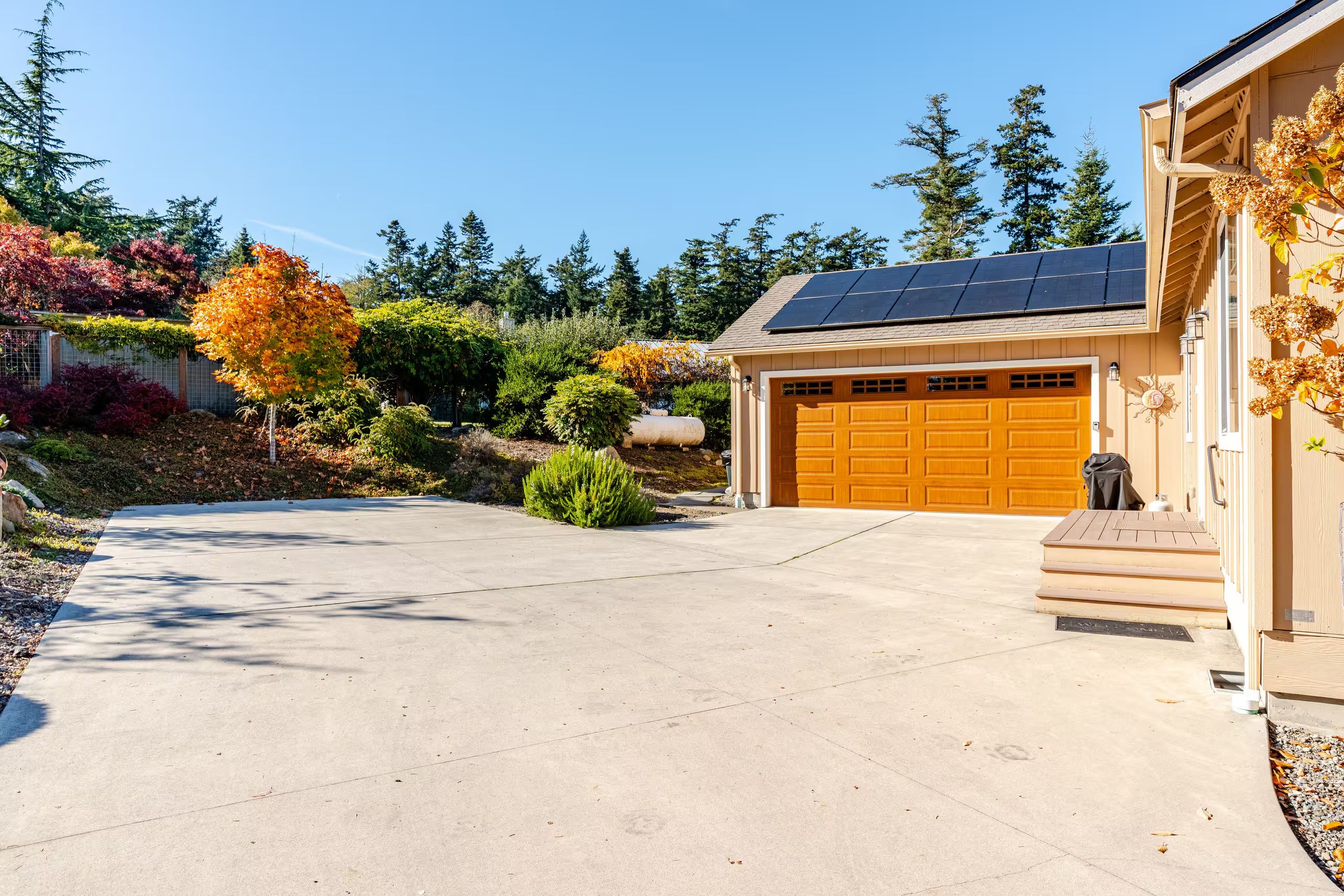 A residential garage with a solar panel roof, a concrete driveway, and a variety of trees and shrubs.