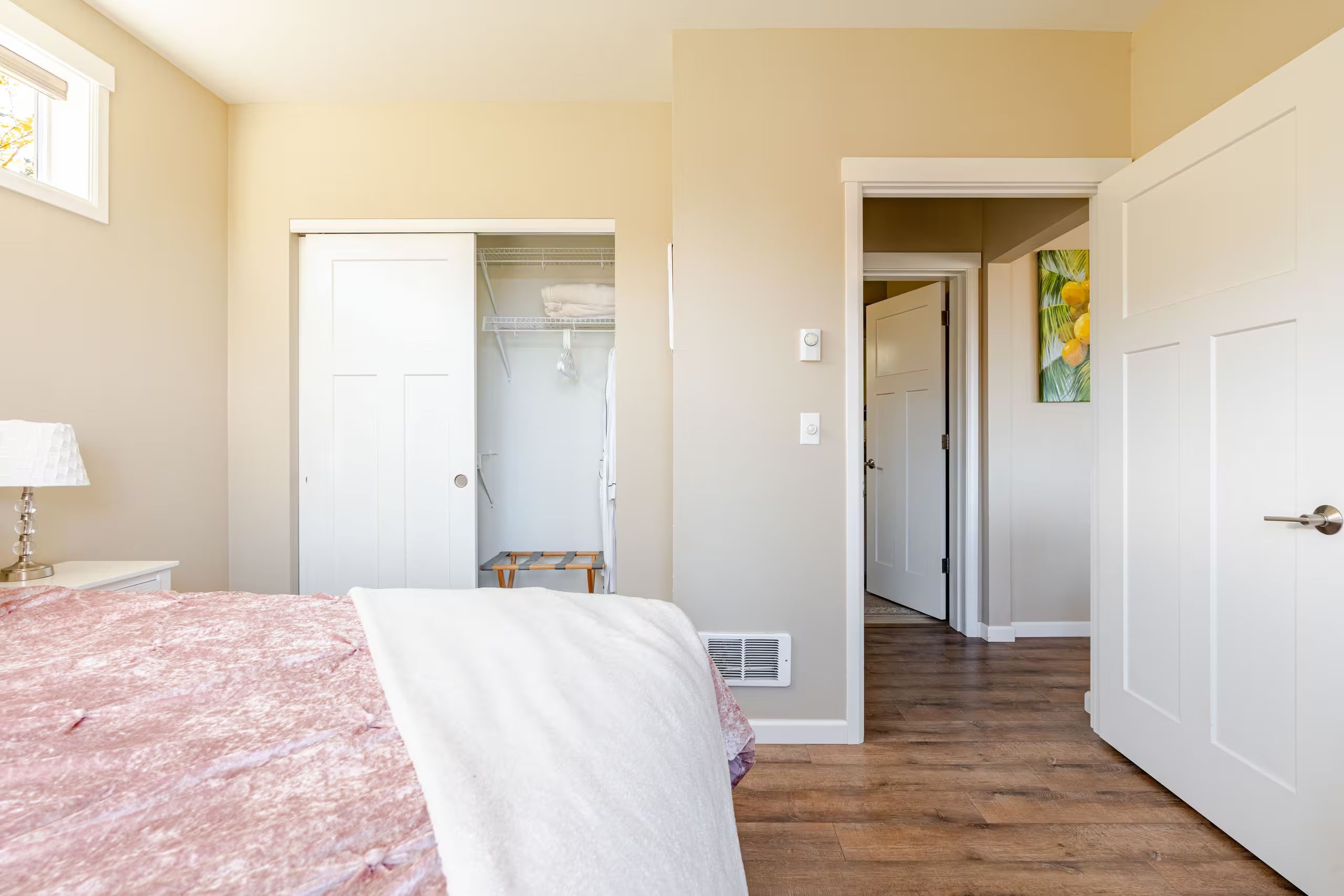 A bedroom with a bed, a nightstand, a lamp, a closet, and a hallway leading to another room.