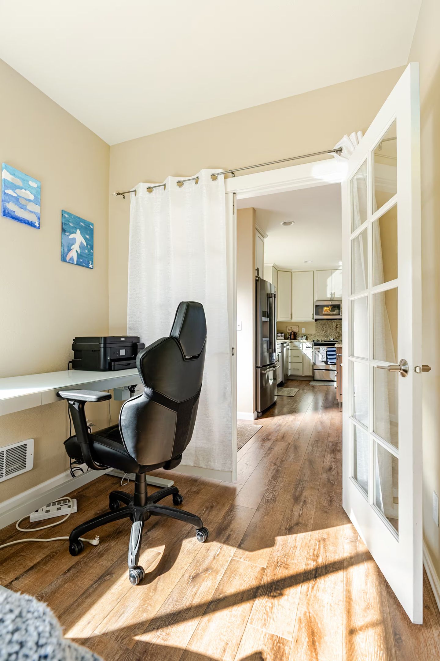 A room with a wooden floor, a black office chair, a desk with a printer, and a view into a kitchen area through an open door.