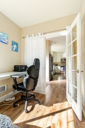 A room with a wooden floor, a black office chair, a desk with a printer, and a view into a kitchen area through an open door.