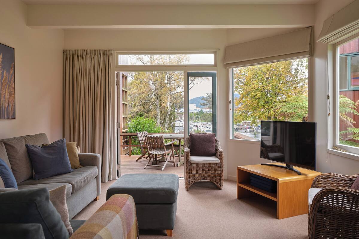 A cozy living room with a sofa, ottoman, wicker chairs, a TV stand, and a view of a marina through large windows.
