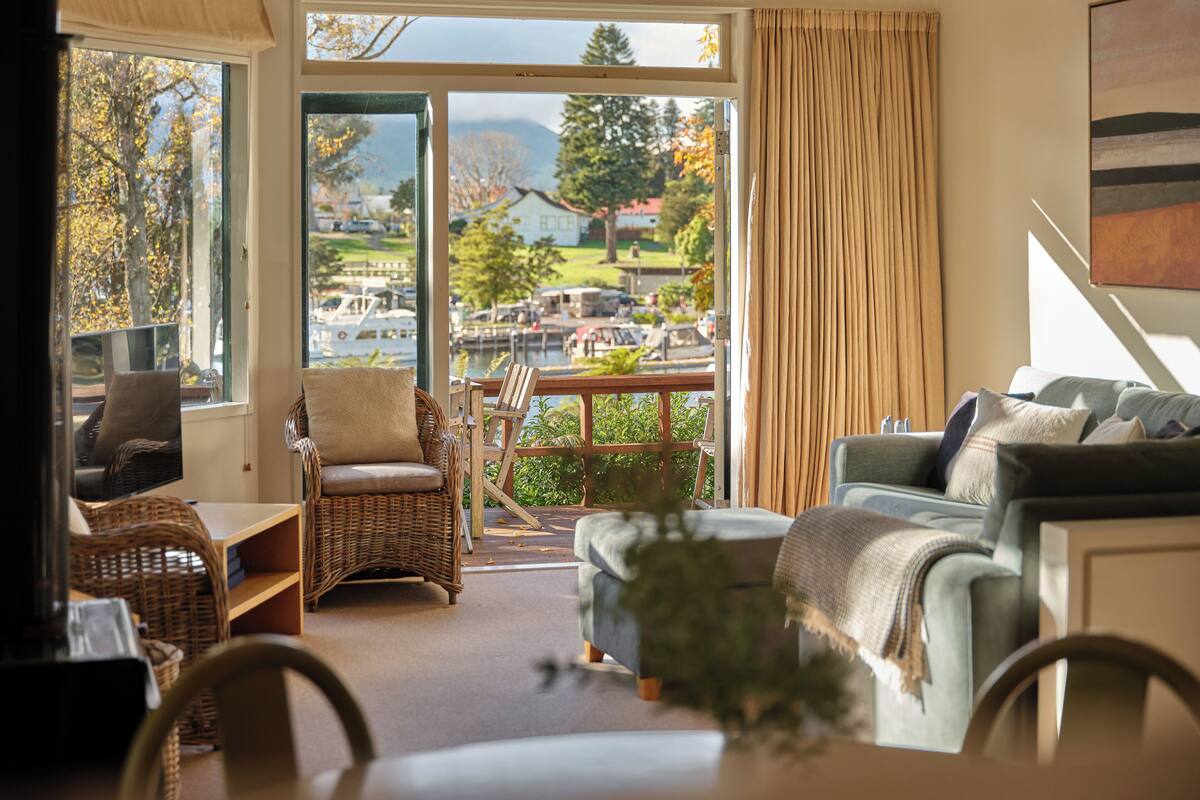 A spacious living room with a view of a marina and mountains. It includes wicker furniture, a sofa, and a dining table.