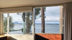 A room with a large window overlooking a body of water and trees, a bed, and a wooden wall.
