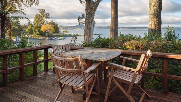 A wooden deck with a table and chairs, overlooking a body of water with boats and a distant cityscape.