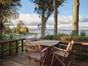 A wooden deck with a table and chairs, overlooking a body of water with boats and a distant cityscape. - The Retreat (Taupo)