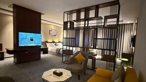 A modern living room with a yellow sofa, a coffee table, and a TV displaying a crowd scene. - Belvedere Hotel (Madinah)