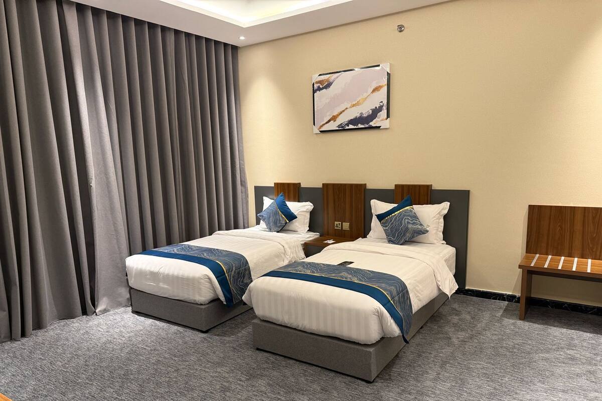 A hotel room with two beds, grey curtains, a wooden bench, and a painting on the wall.