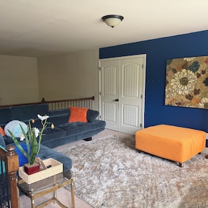 Un salon avec un canapé bleu, un pouf orange, une table basse en bois et un tableau floral accroché au mur.