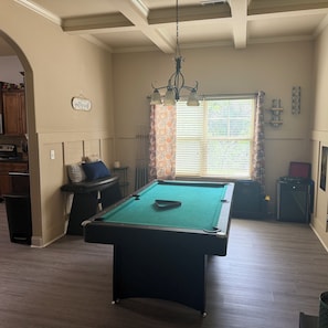 Une table de billard dans une pièce dotée d’un plancher en bois, d’un ventilateur de plafond et d’une fenêtre avec des stores.