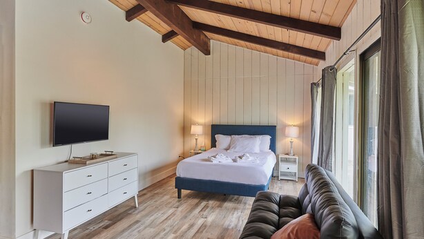A bedroom with a wooden ceiling, a bed with white bedding, a TV, a white dresser, a brown leather sofa, and a window with curtains.