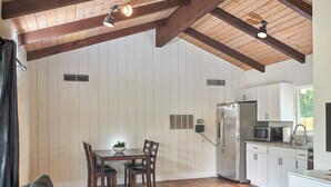A modern kitchen with white cabinets, a stainless steel refrigerator, and a microwave. There is a dining table with chairs and a bowl of fruit. The room has wooden floors and a ceiling with exposed beams.