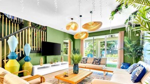 Un salon moderne avec un mur d’accent vert, un téléviseur à écran plat, des meubles en osier et une grande fenêtre donnant sur l’extérieur.