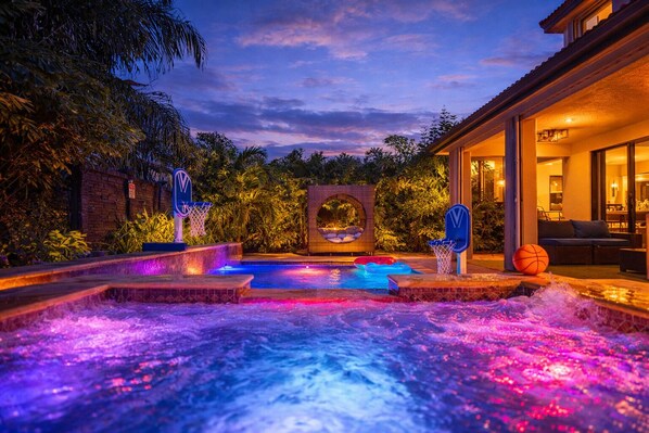 A heated outdoor pool with two basketball hoops, surrounded by lush greenery and a brick wall.