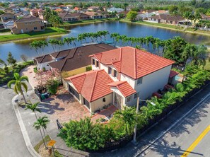 A residential area with a house featuring a red-tiled roof, surrounded by a small pond and palm trees.