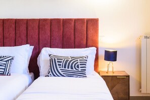 A hotel room with two beds, a red upholstered headboard, a bedside lamp, and a radiator.