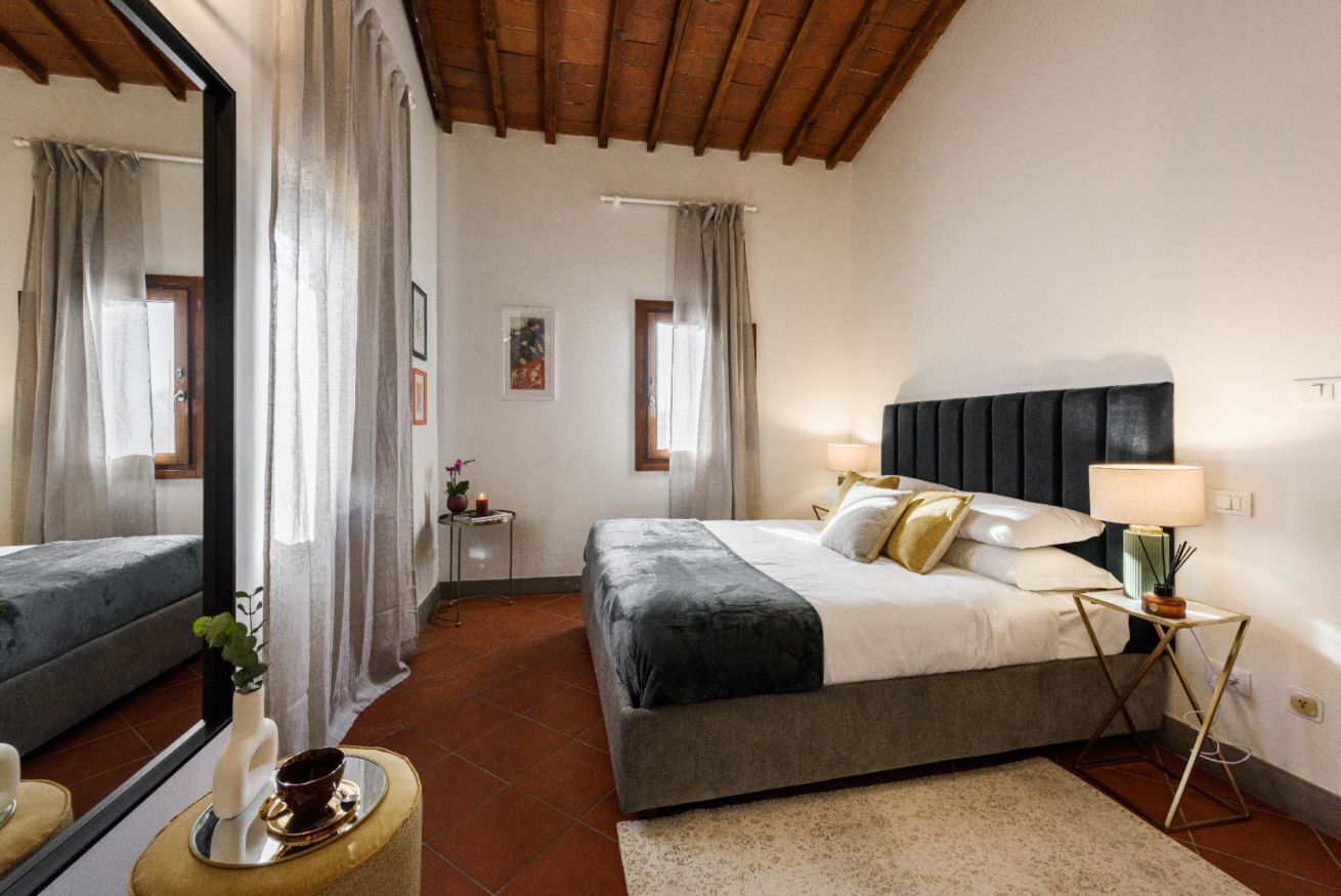 A bedroom with a large bed, a bedside table with a lamp, a mirror, and a view of the outside.